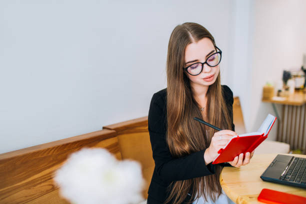 Young attractive businesswoman in glasses writing in a red notepad, sitting in front of the laptop in a cafe.